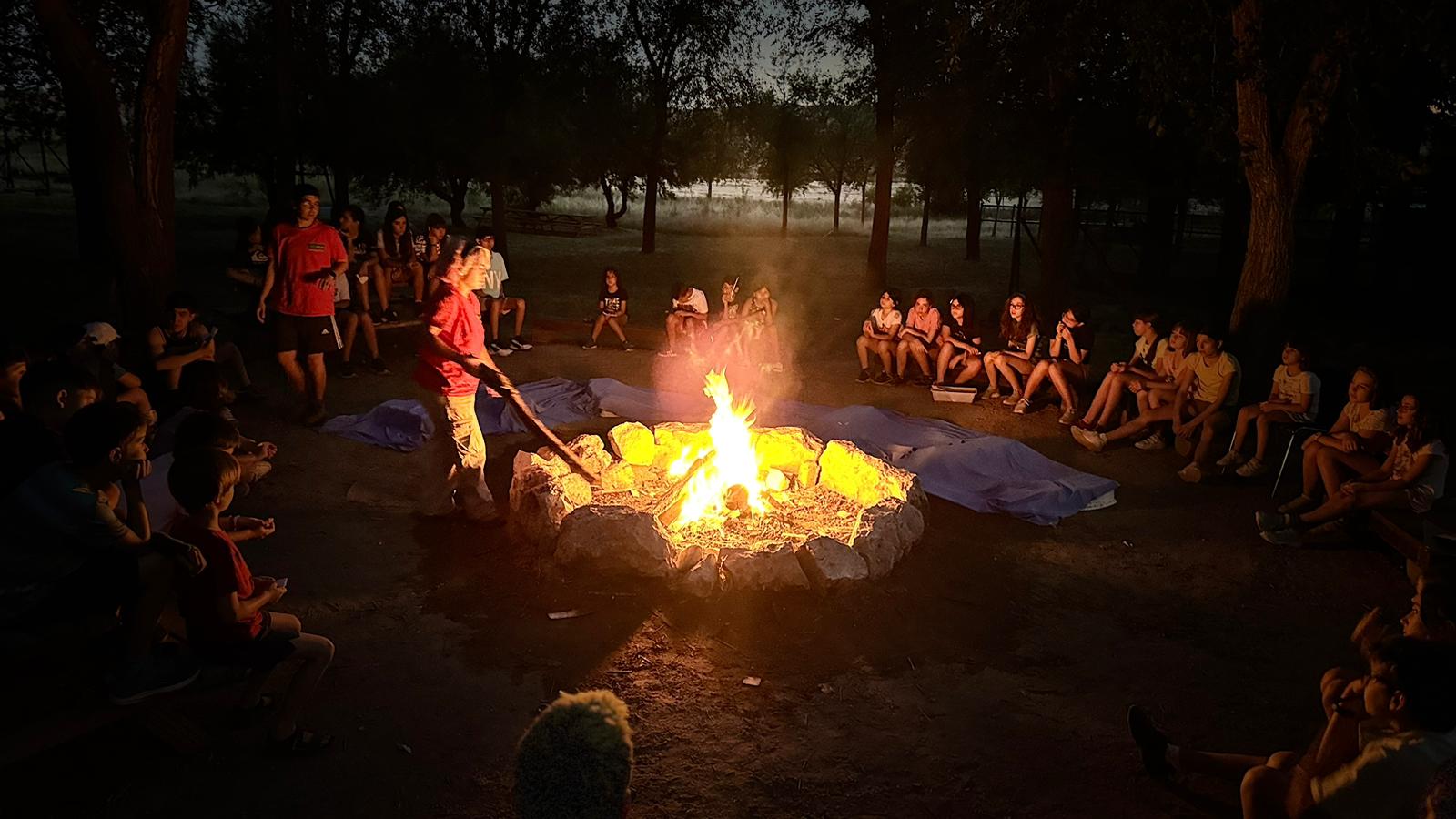 Un amplio grupo de niños y niñas sentados en el suelo por la noche, al rededor de una hoguera, en el campamento de verano NF